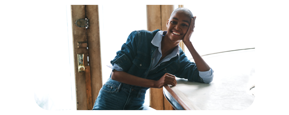 Smiling woman wearing a denim jacket and jeans leaning on a counter.