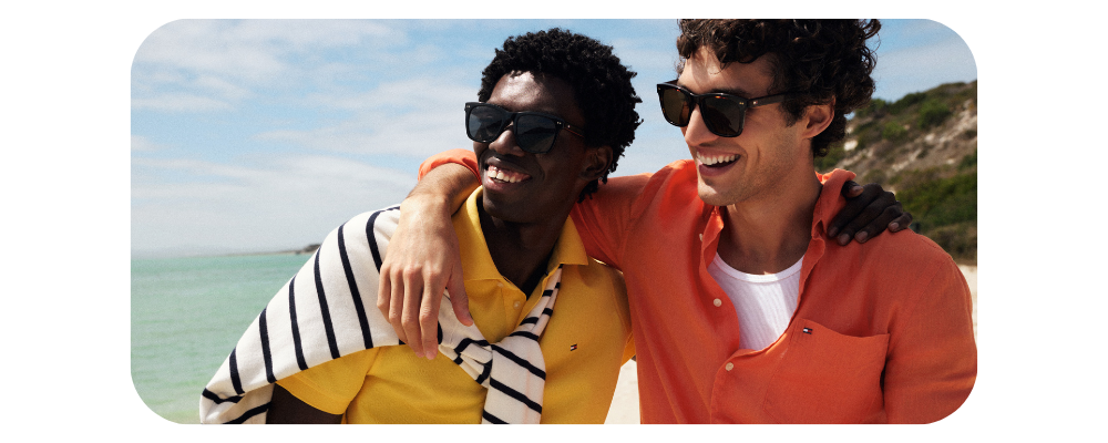 Two smiling men in sunglasses wearing summer shirts posing outdoors by the beach.