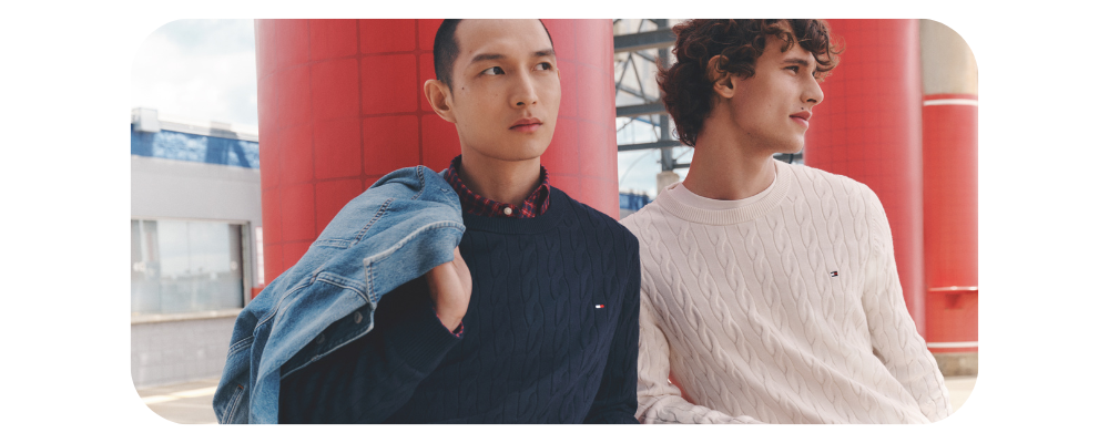 Two men wearing cable-knit sweaters in navy and cream against a red architectural background.