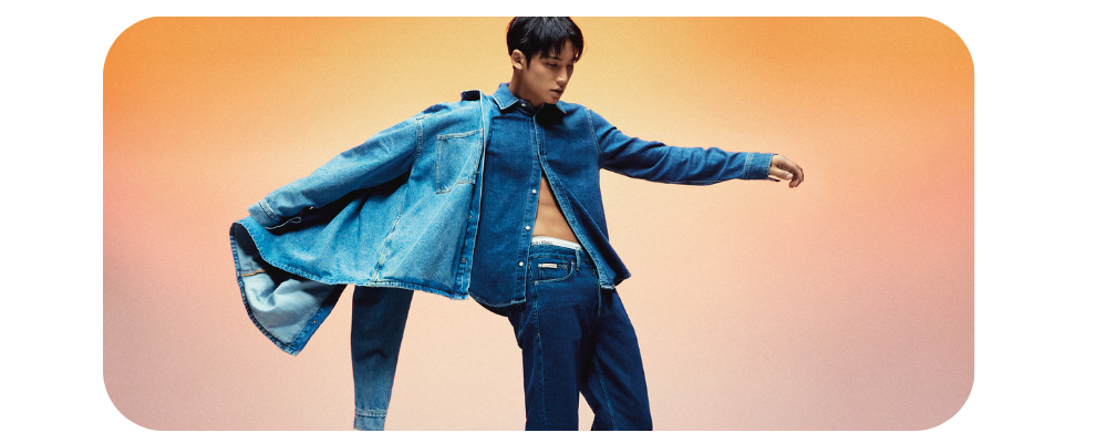 Man posing in a denim jacket and matching jeans against a gradient orange background.