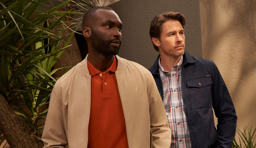 Two men wearing a burnt orange polo shirt under a beige jacket and a navy blue utility jacket.
