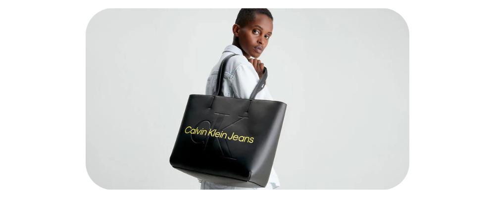 Woman wearing a white denim jacket carrying a black Calvin Klein Jeans tote bag.